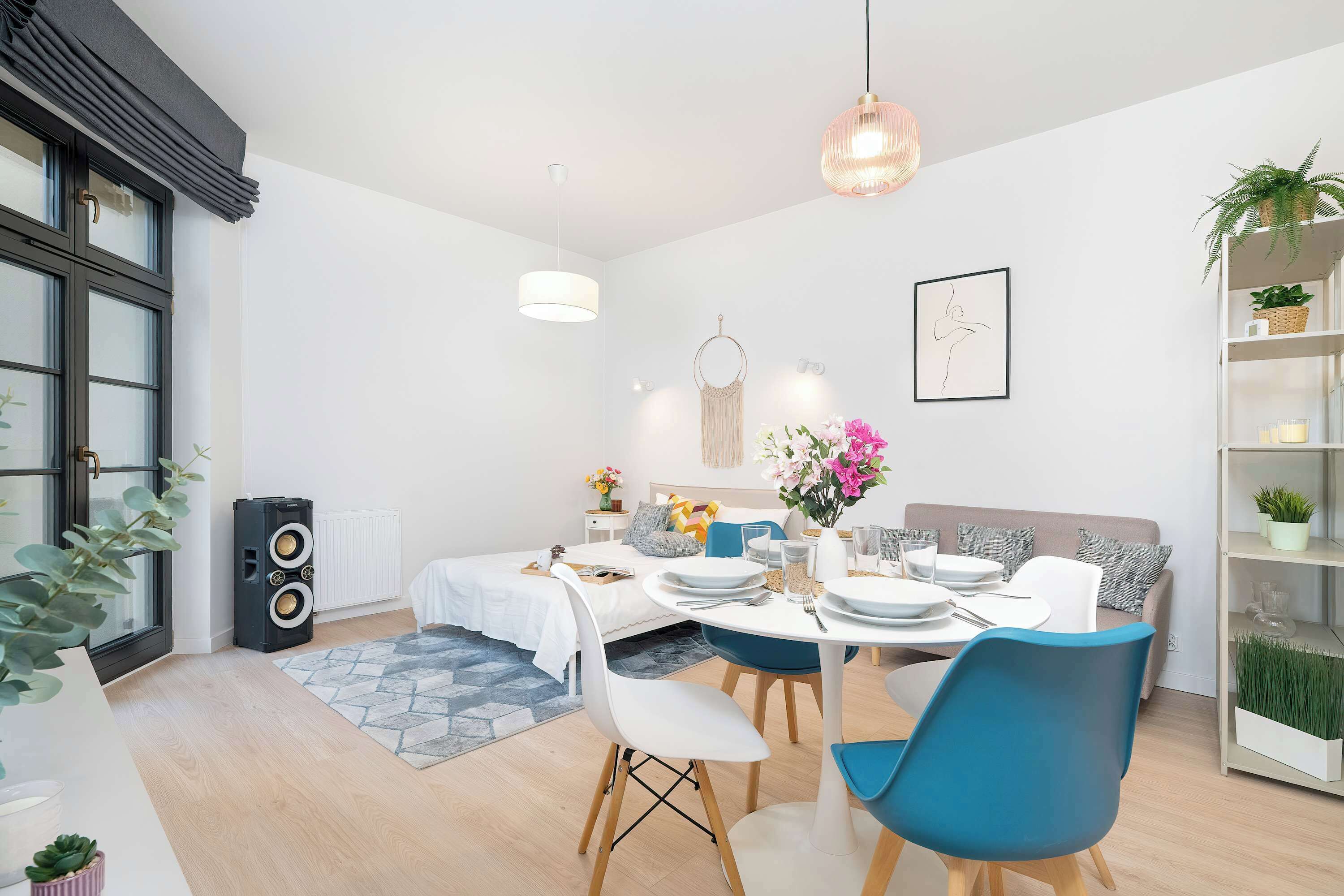 A modern dining area with a round table set for four, a blue chair, a white sofa, and a bookshelf with plants.