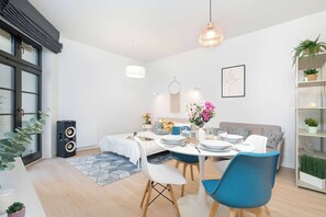 A modern dining area with a round table set for four, a blue chair, a white sofa, and a bookshelf with plants. - Stylish Apartment Near the Old Market Square | Pet Friendly (Poznań)