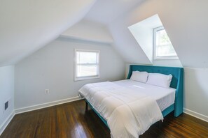 Un dormitorio con techo inclinado, una cama con ropa blanca, cabecera color turquesa y una ventana con persianas.