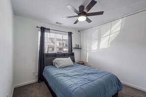 Een slaapkamer met een bed, een plafondventilator, een raam met gordijnen en uitzicht op gebouwen.