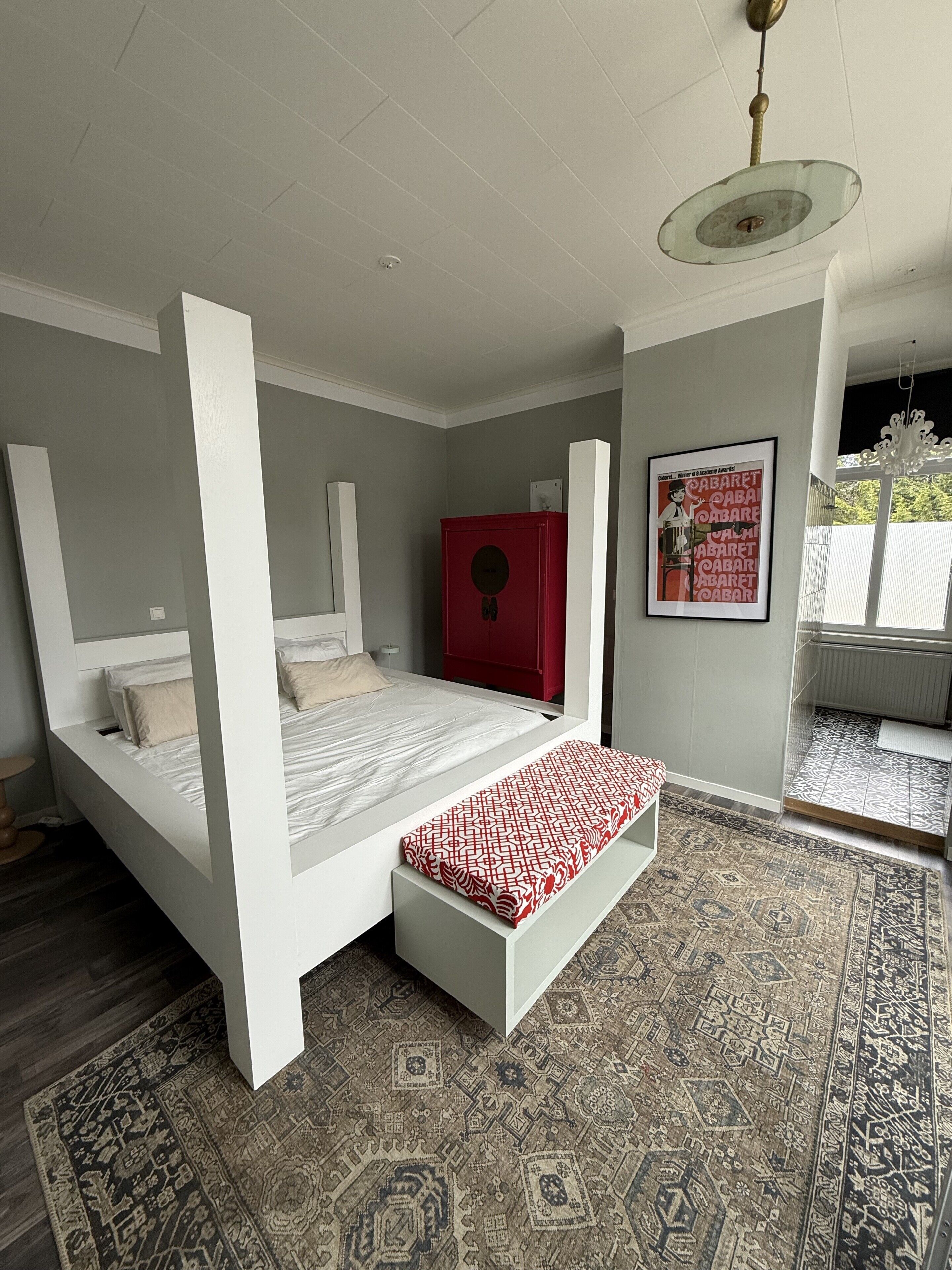 Een slaapkamer met een hemelbed, een rode kast, een kamerkleed met patroon en een ingelijste foto aan de muur.