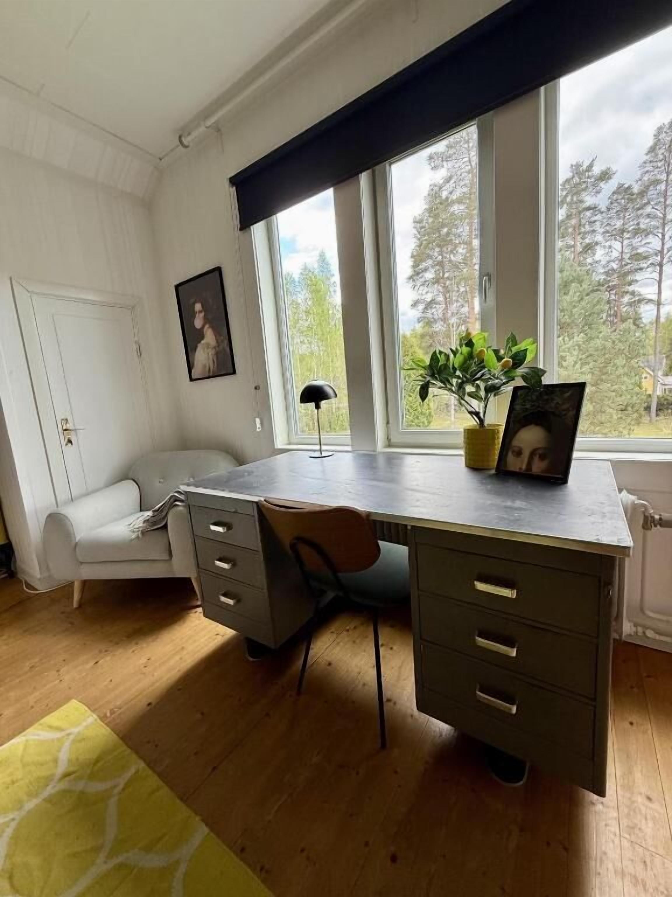 A room with a wooden desk, a chair, a white sofa, and a window with a view of trees.