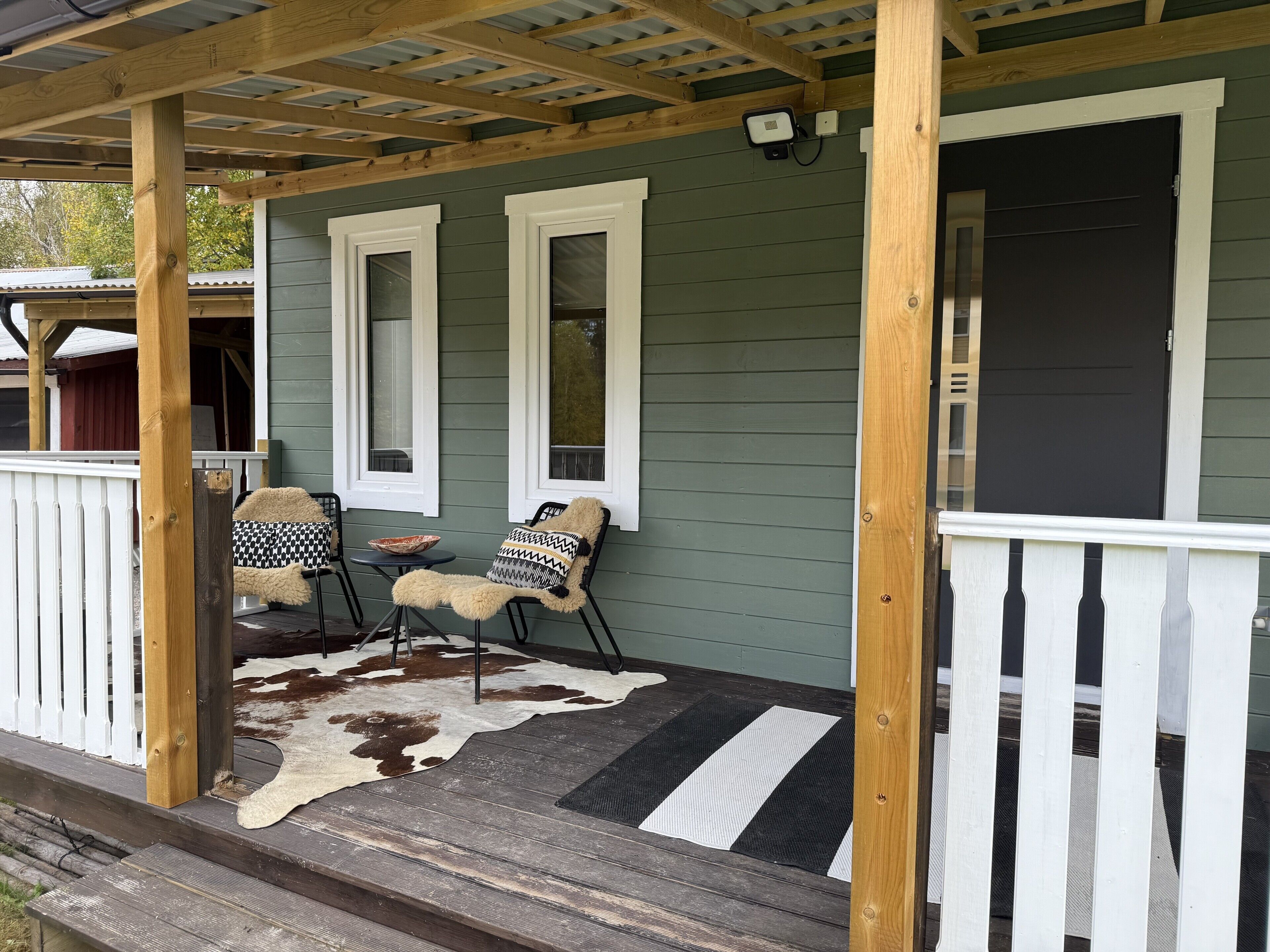 Eine Veranda mit Holzgeländer, zwei Stühlen, einer Kuhfelldecke und einer schwarzen Tür.