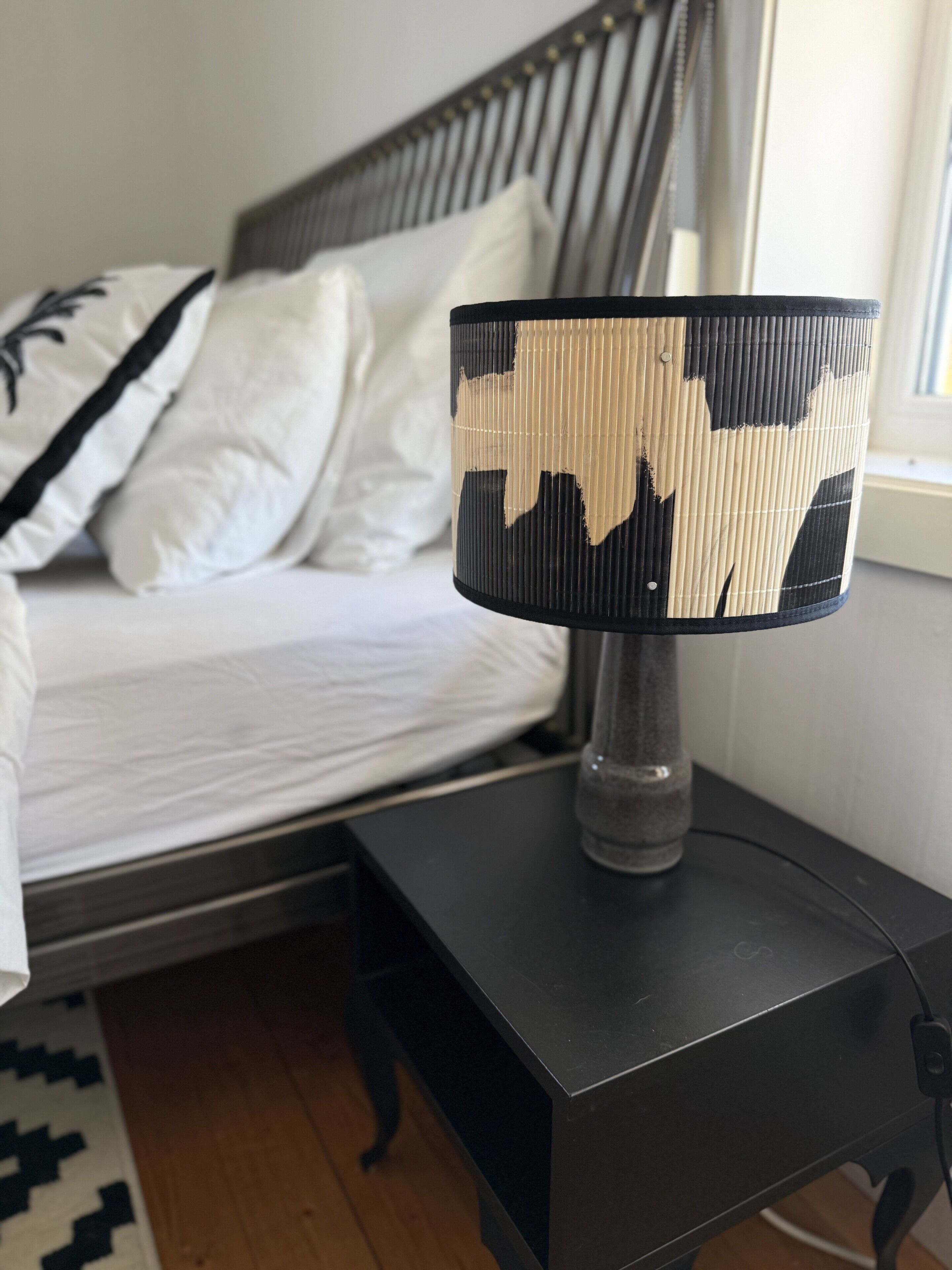 Una cama con ropa de cama blanca, una mesita de noche con una lámpara y una ventana con cortinas.