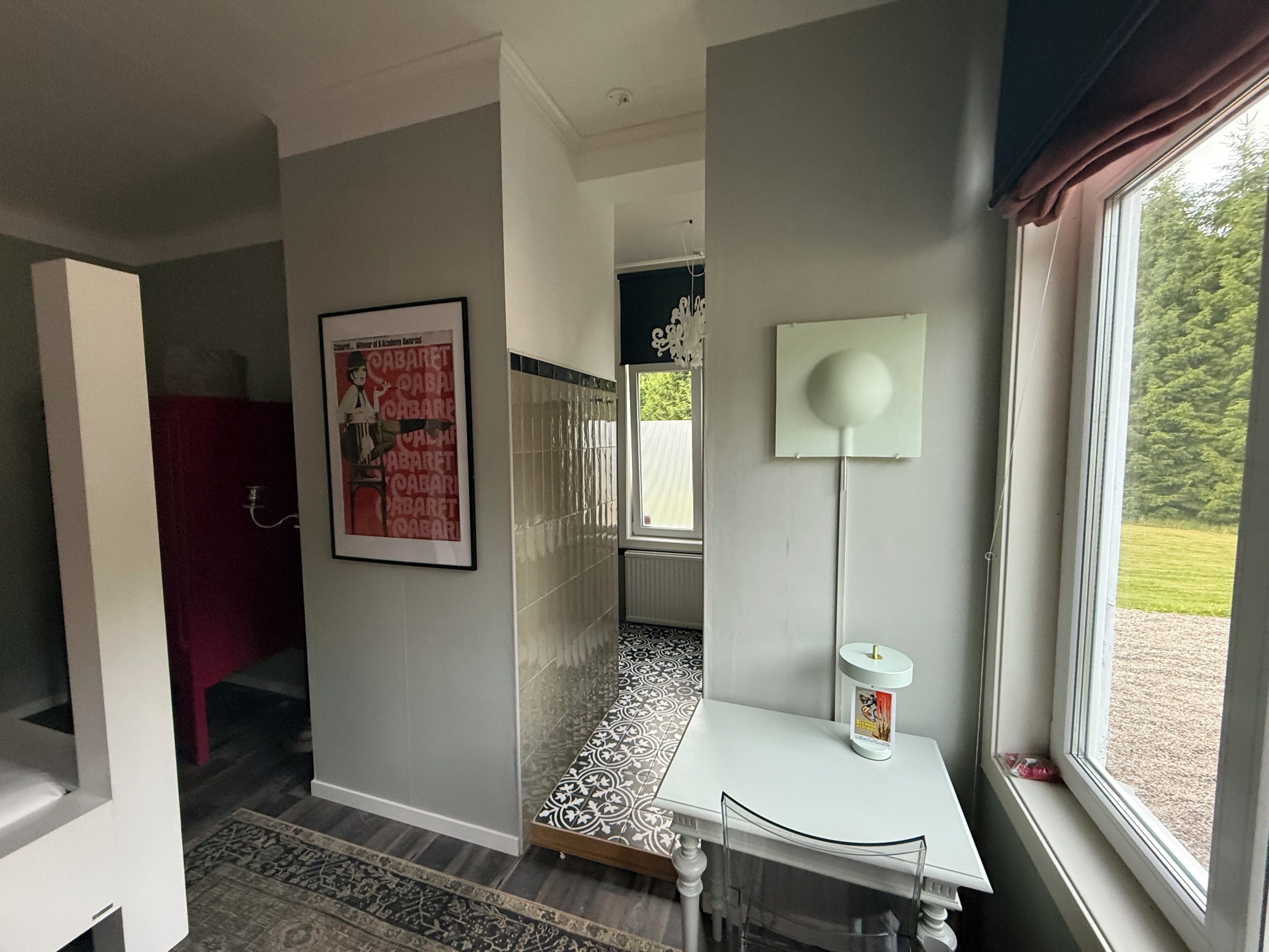 Une chambre d’hôtel moderne avec vue sur l’extérieur, une table blanche avec une lampe, un tapis à motifs et une affiche accrochée au mur.