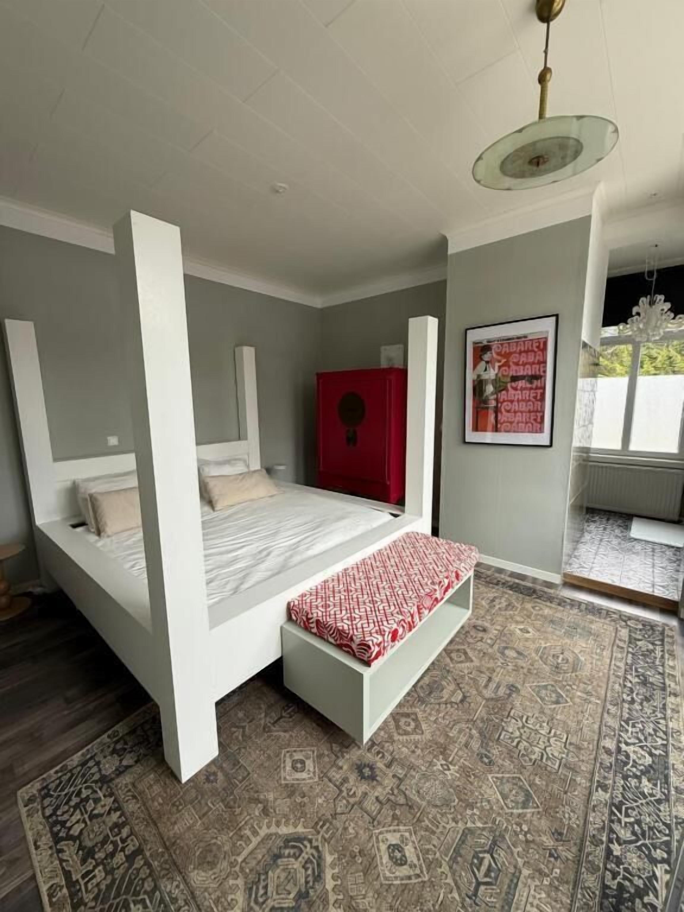 Une chambre à coucher avec un lit, une armoire rouge, un tapis à motifs et un tableau encadré accroché au mur.