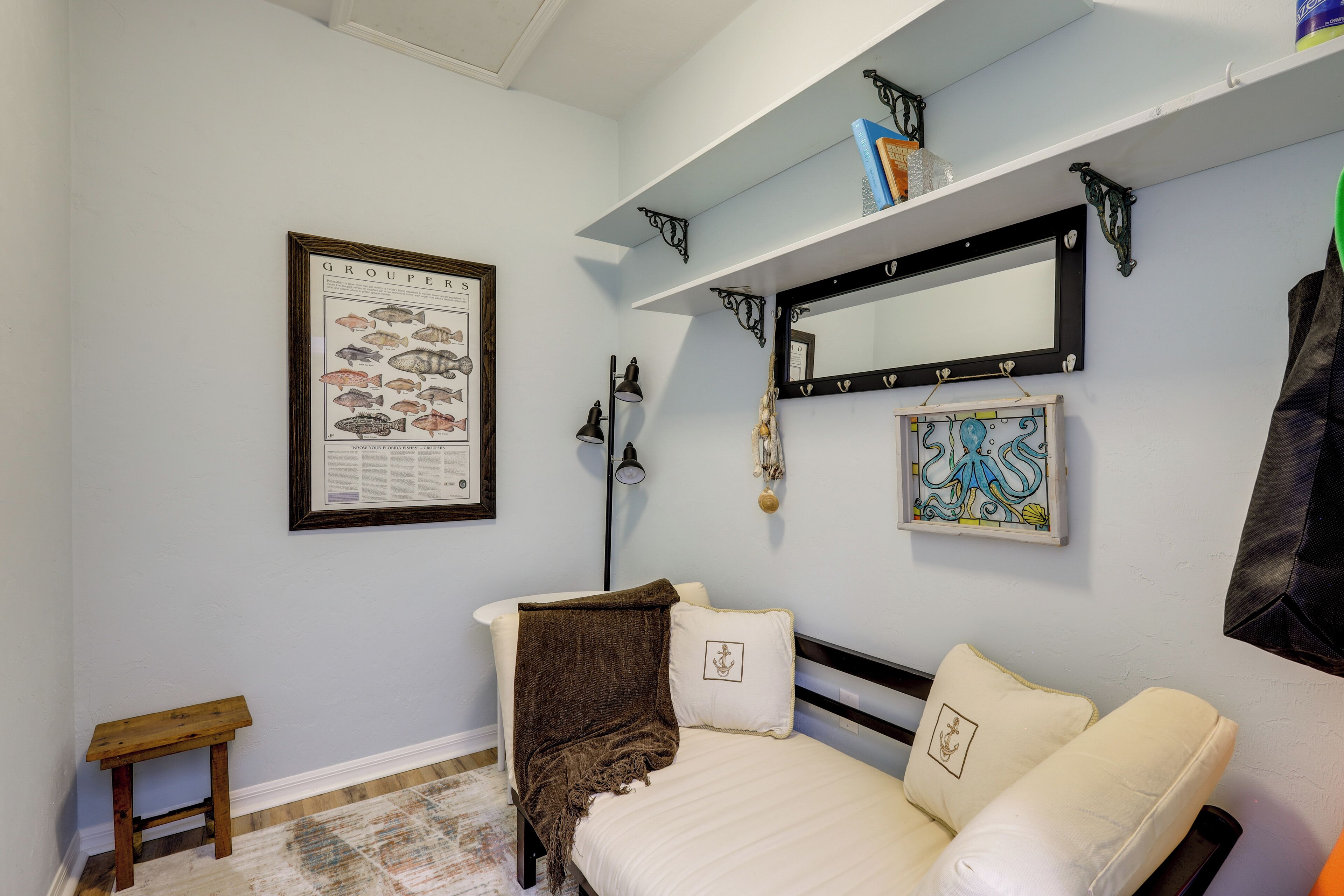 A bedroom with a bed, a small bedside table, a framed fish chart, a mirror, and a shelf with books and decorative items.