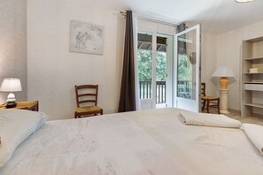 A hotel room with a bed, a chair, a table, a lamp, a picture on the wall, and a balcony with a view of trees.