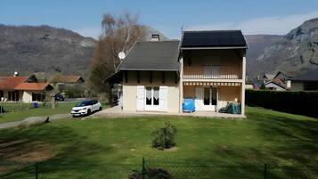Une maison à deux étages avec un balcon, une voiture garée devant et un paysage montagneux en arrière-plan.