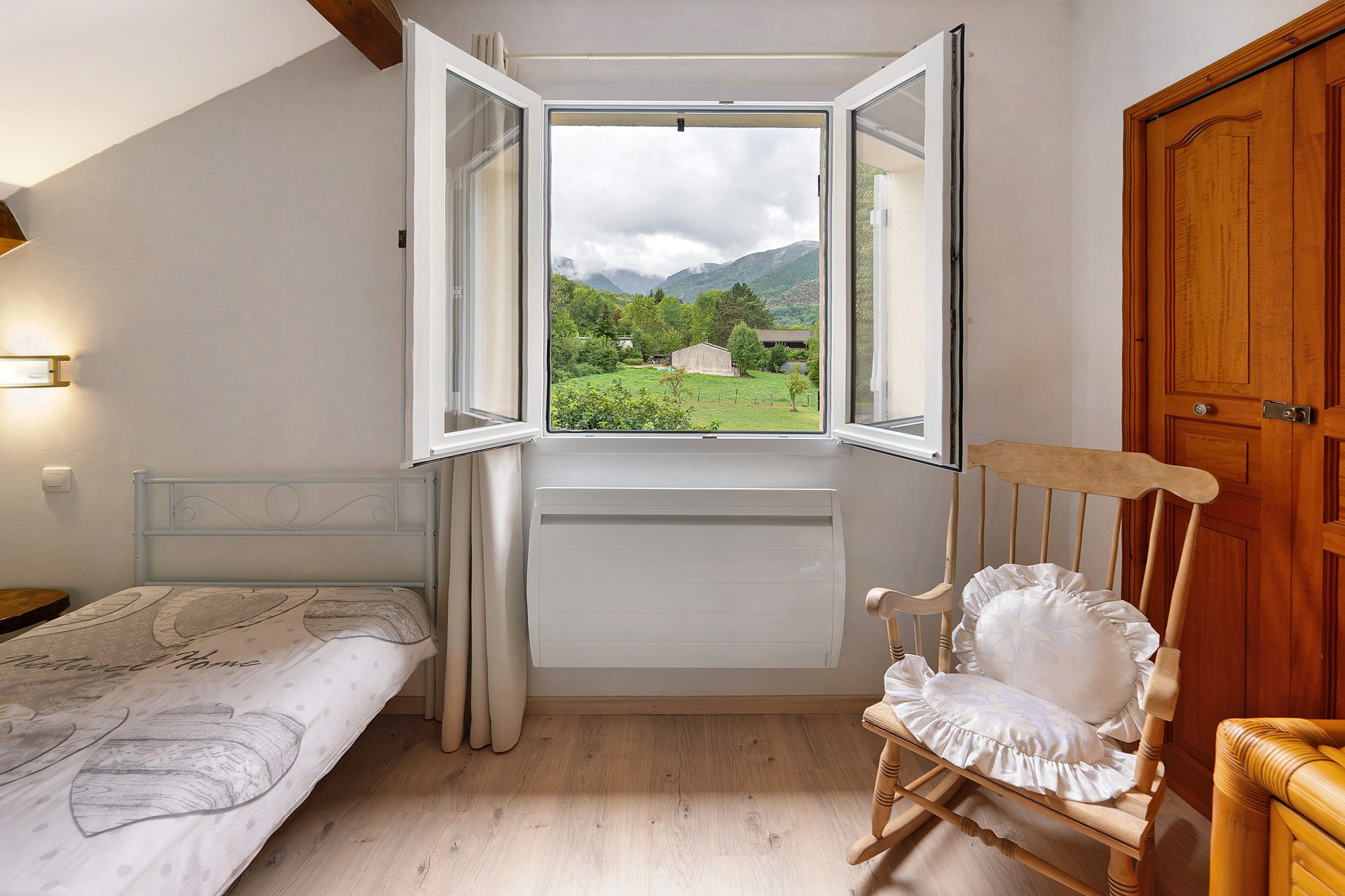 A bedroom with a bed, a wooden chair, a window with an open shutter, and a door.