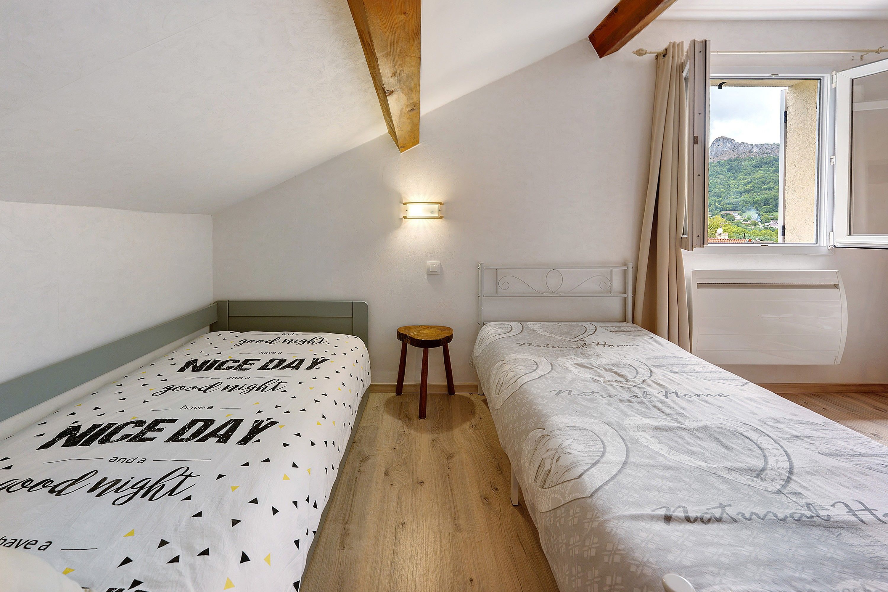 Une chambre avec deux lits, un petit tabouret, une lampe fixée au mur et une fenêtre avec des rideaux.