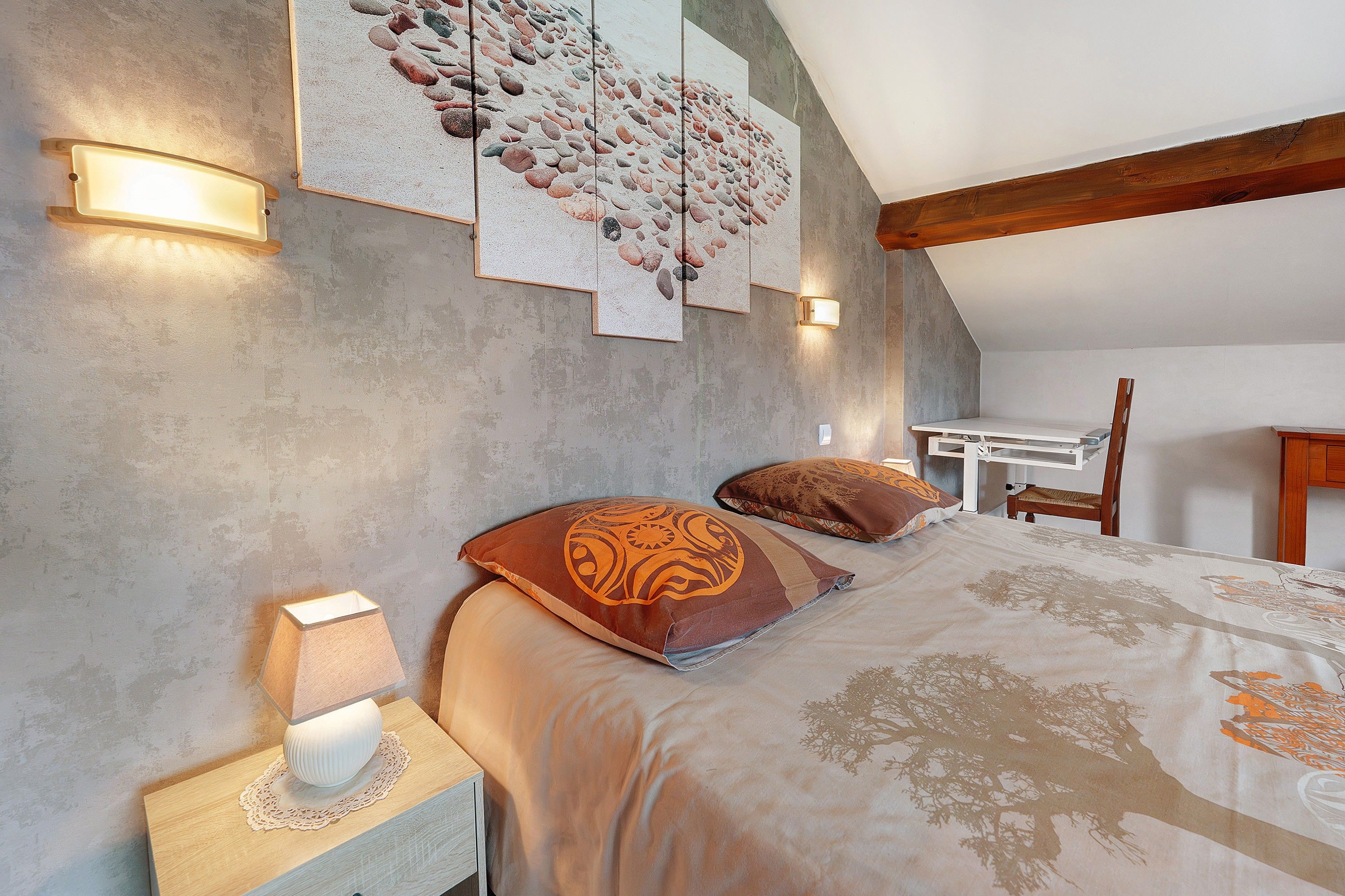 Une chambre avec un plafond en pente, un lit recouvert d’une couvre-lit à motifs d’arbres, deux oreillers ornés de motifs tribaux, une table de chevet avec une lampe et une œuvre d’art murale représentant des galets.