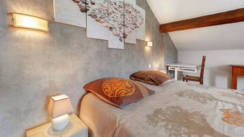 Une chambre avec un plafond en pente, un lit recouvert d’une couvre-lit à motifs d’arbres, deux oreillers ornés de motifs tribaux, une table de chevet avec une lampe et une œuvre d’art murale représentant des galets.