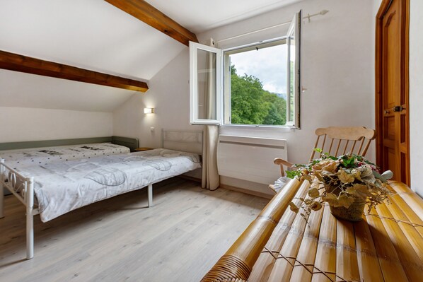 A small, single-room apartment with a sloped ceiling, a bed, a table with a vase of flowers, and a window with a view of greenery.