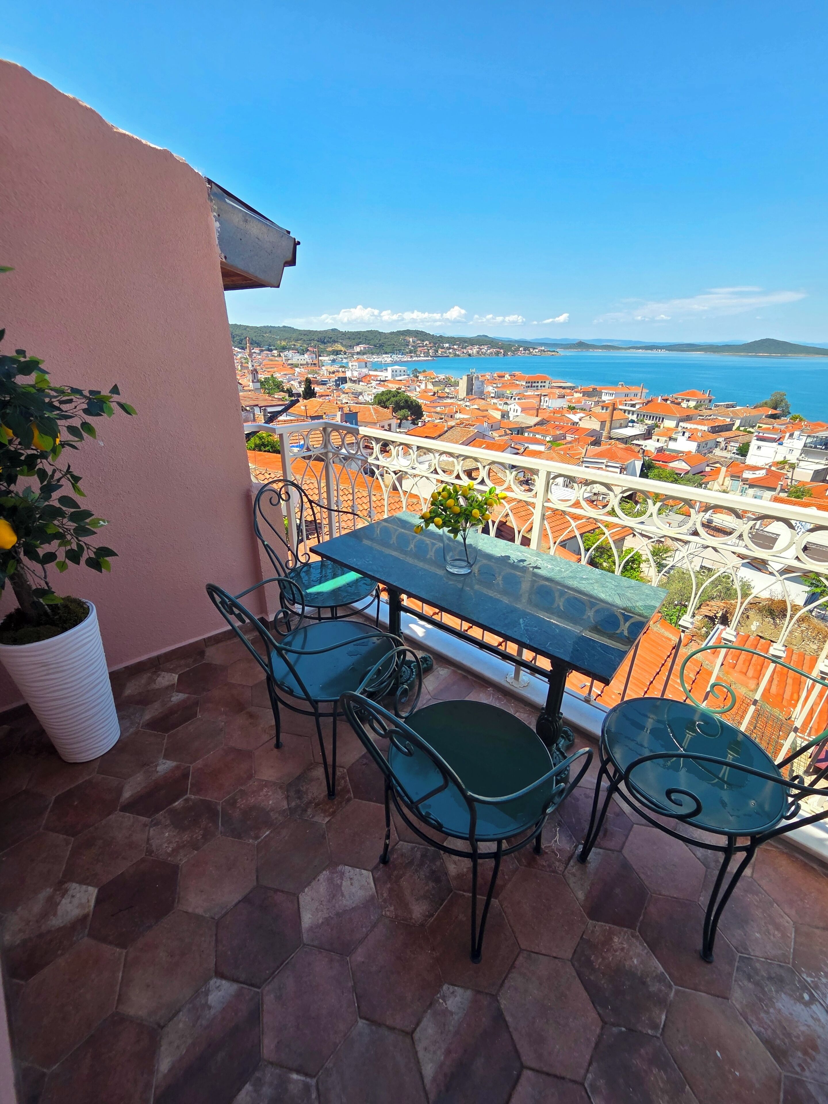 A balcony with a table and chairs, overlooking a coastal town with red-roofed buildings.