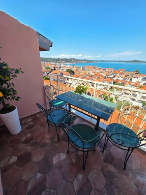 A balcony with a table and chairs, overlooking a coastal town with red-roofed buildings.