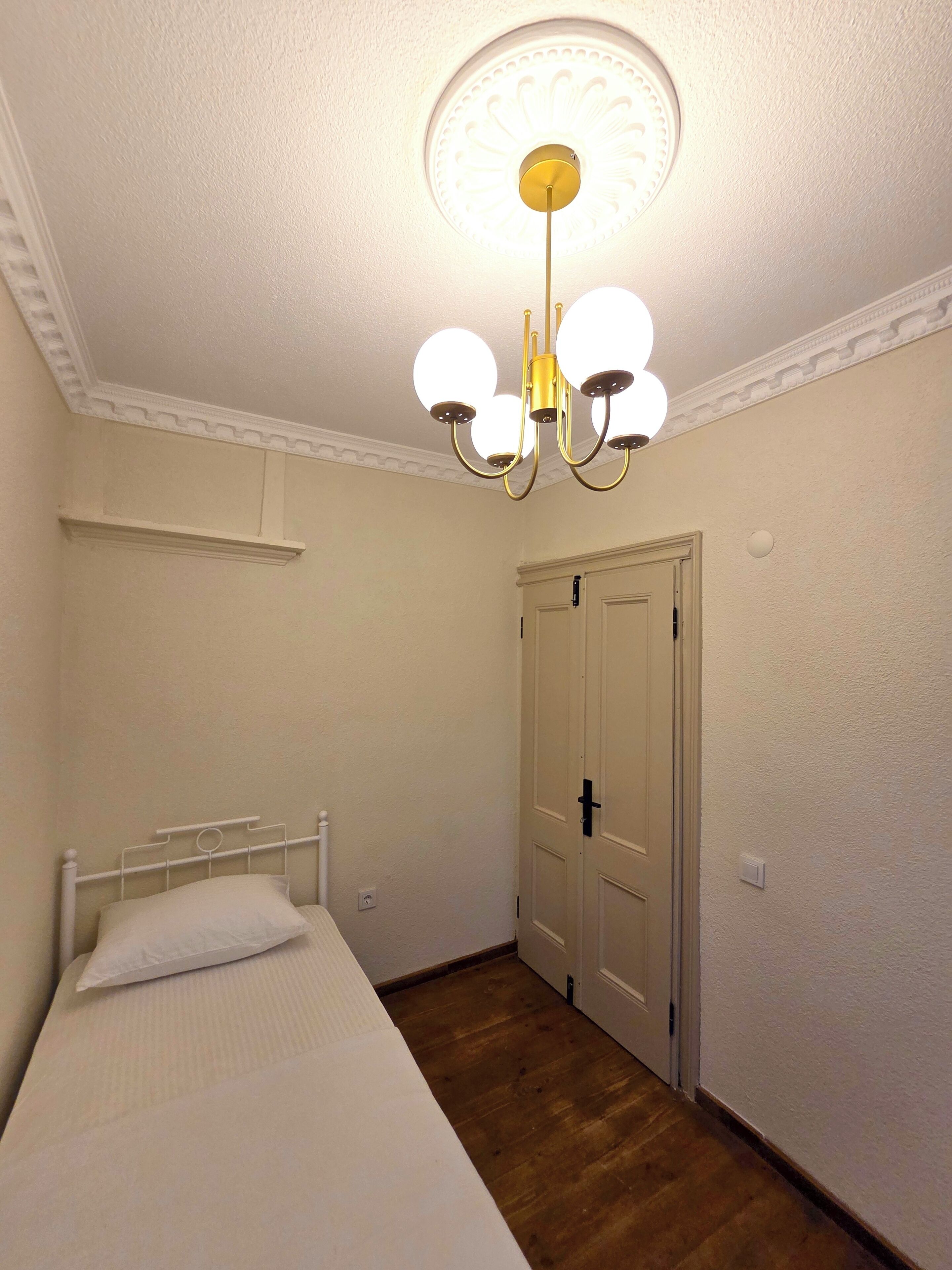 Ein Schlafzimmer mit einem Einzelbett, Holzboden, weißer Tür und einer Deckenleuchte.