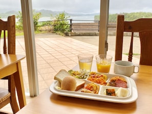 A tray with a sandwich, pasta, and a drink, placed on a table with a view of a body of water and mountains.