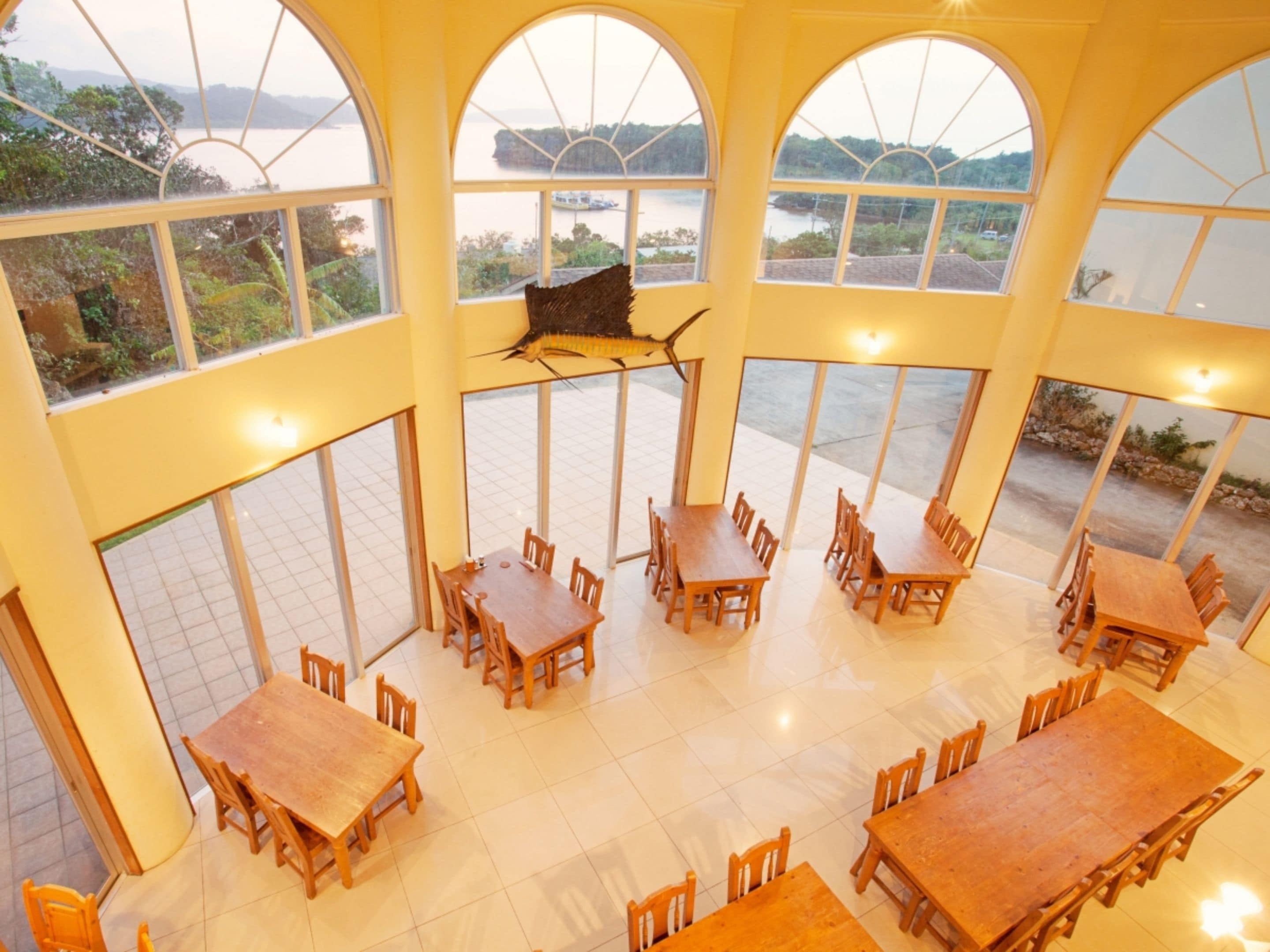 A dining area with multiple tables and chairs, large windows with arched tops, and a view of a body of water and greenery outside.