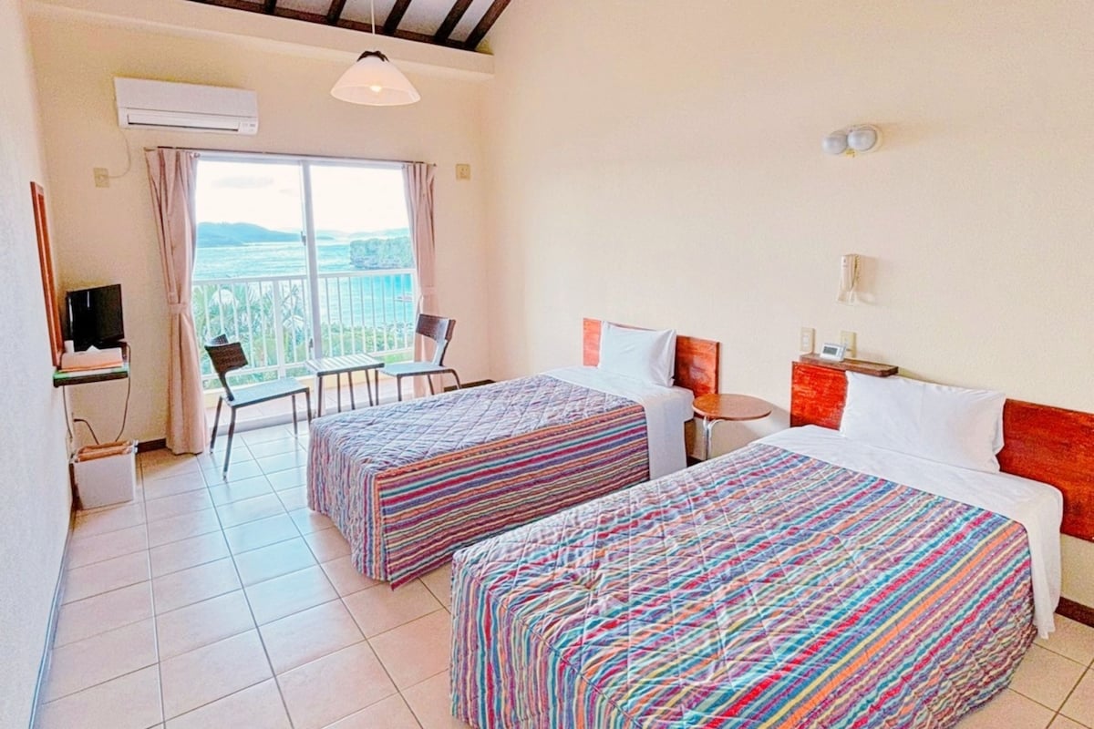 A hotel room with two beds, a desk with a computer, a chair, a small table, and a window with a view.