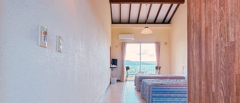A hotel room with a tiled floor, a bed with a striped bedspread, a desk with a chair, and a window with curtains.