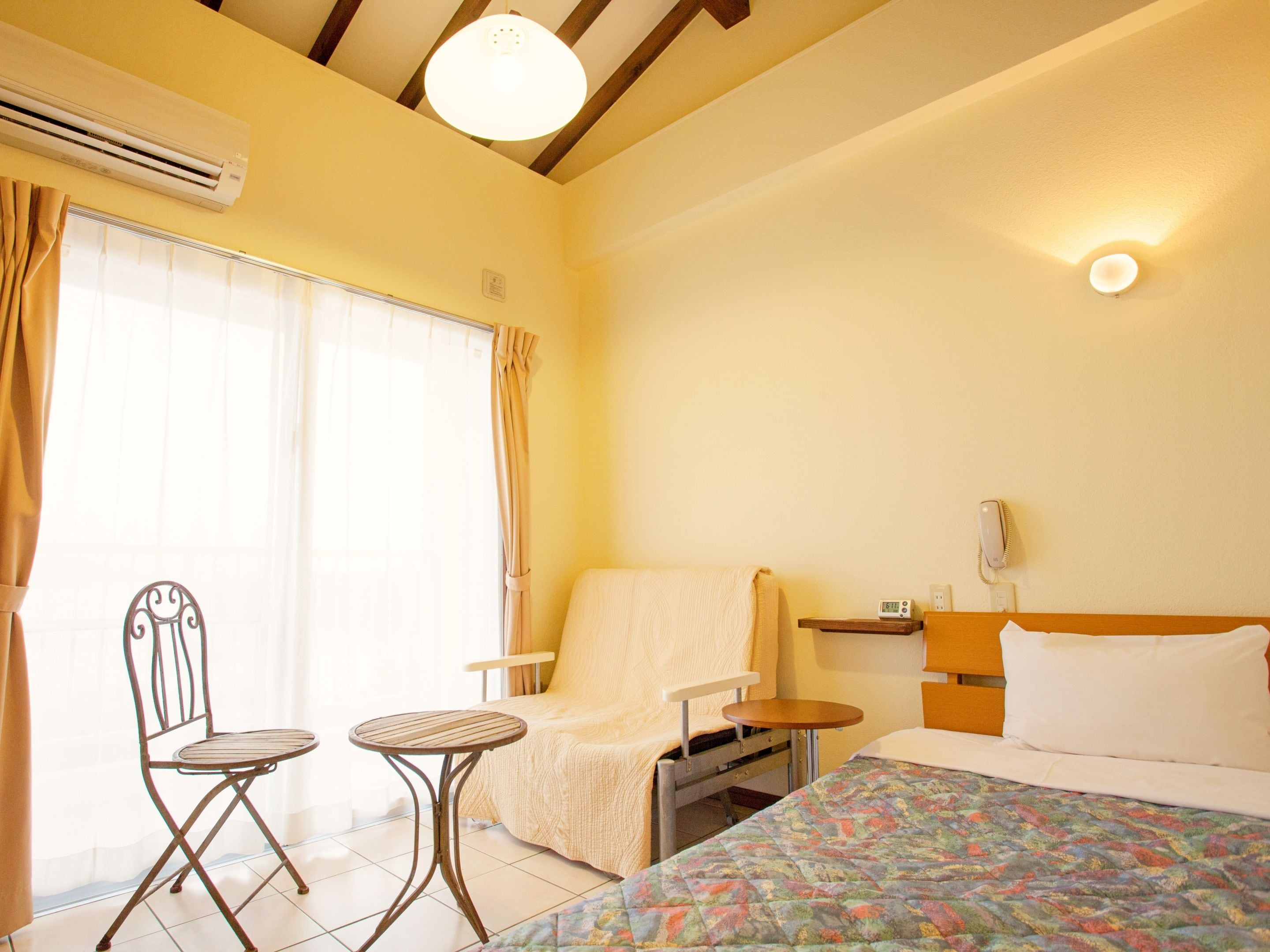 A hotel room with a bed, a chair, a small table, an air conditioning unit, and a bedside table with a lamp.