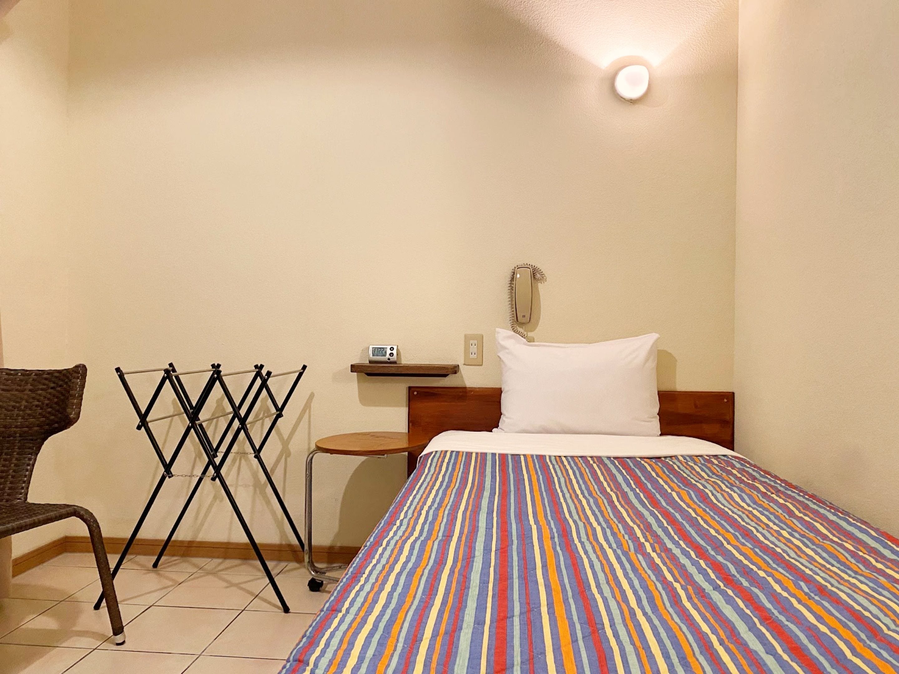 A hotel room with a bed, a chair, a drying rack, a bedside table with a lamp, and a wall-mounted phone.