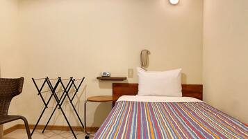 A hotel room with a bed, a chair, a drying rack, a bedside table with a lamp, and a wall-mounted phone.