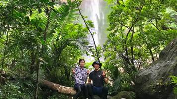 Two individuals seated on rocks surrounded by lush greenery and a waterfall in the background.