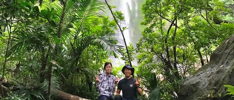 Two individuals seated on rocks surrounded by lush greenery and a waterfall in the background.