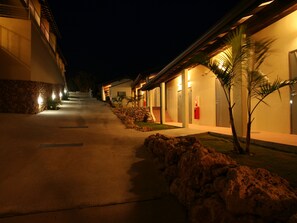 A nighttime view of a hotel corridor with multiple rooms, a stone wall on one side, and a palm tree on the other. - HOTEL ILMARE UNARIZAKI (Taketomi)