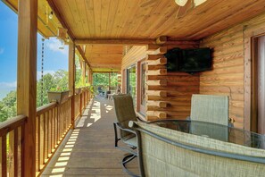 Una terraza de madera con una mesa y sillas de vidrio, un televisor montado en la pared y vistas al exterior.