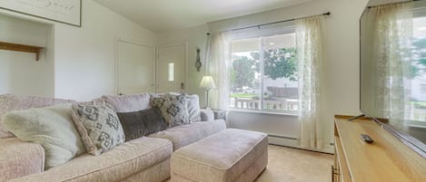 Ett vardagsrum med en hörnsoffa, en liten pall, en platt-TV och ett fönster med gardiner.