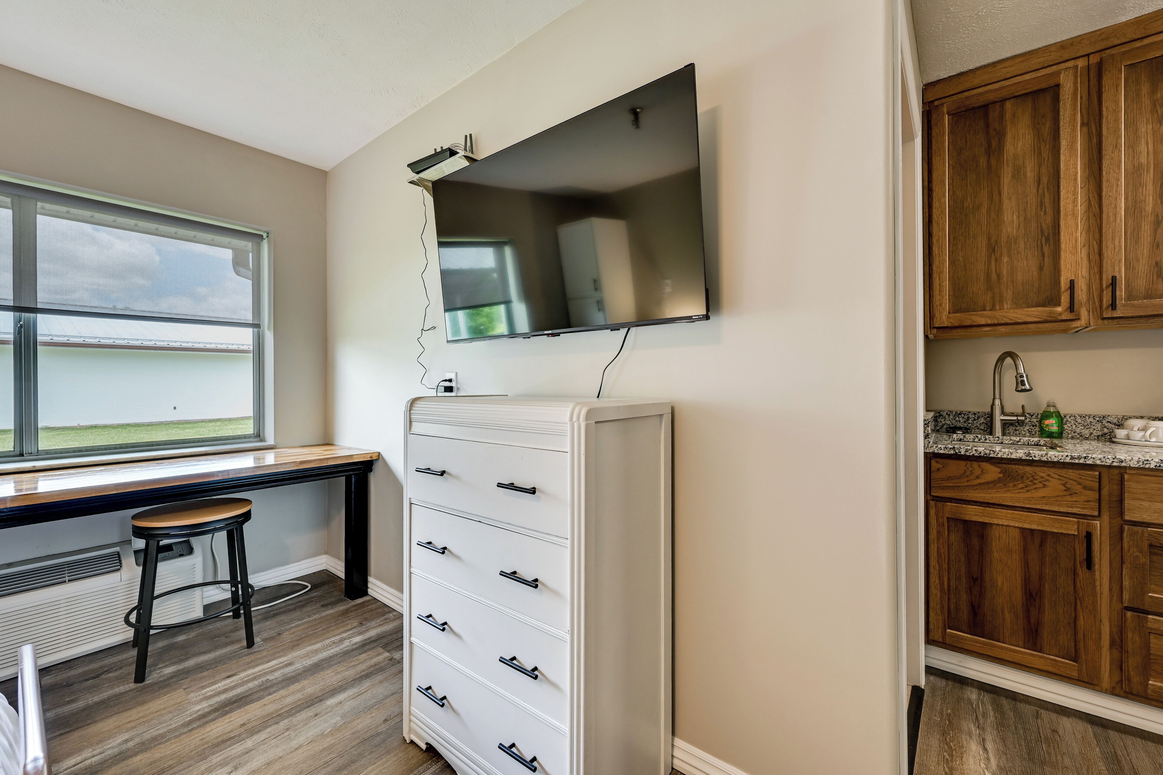 A room with a desk, a white filing cabinet, a television mounted on the wall, and wooden cabinets.