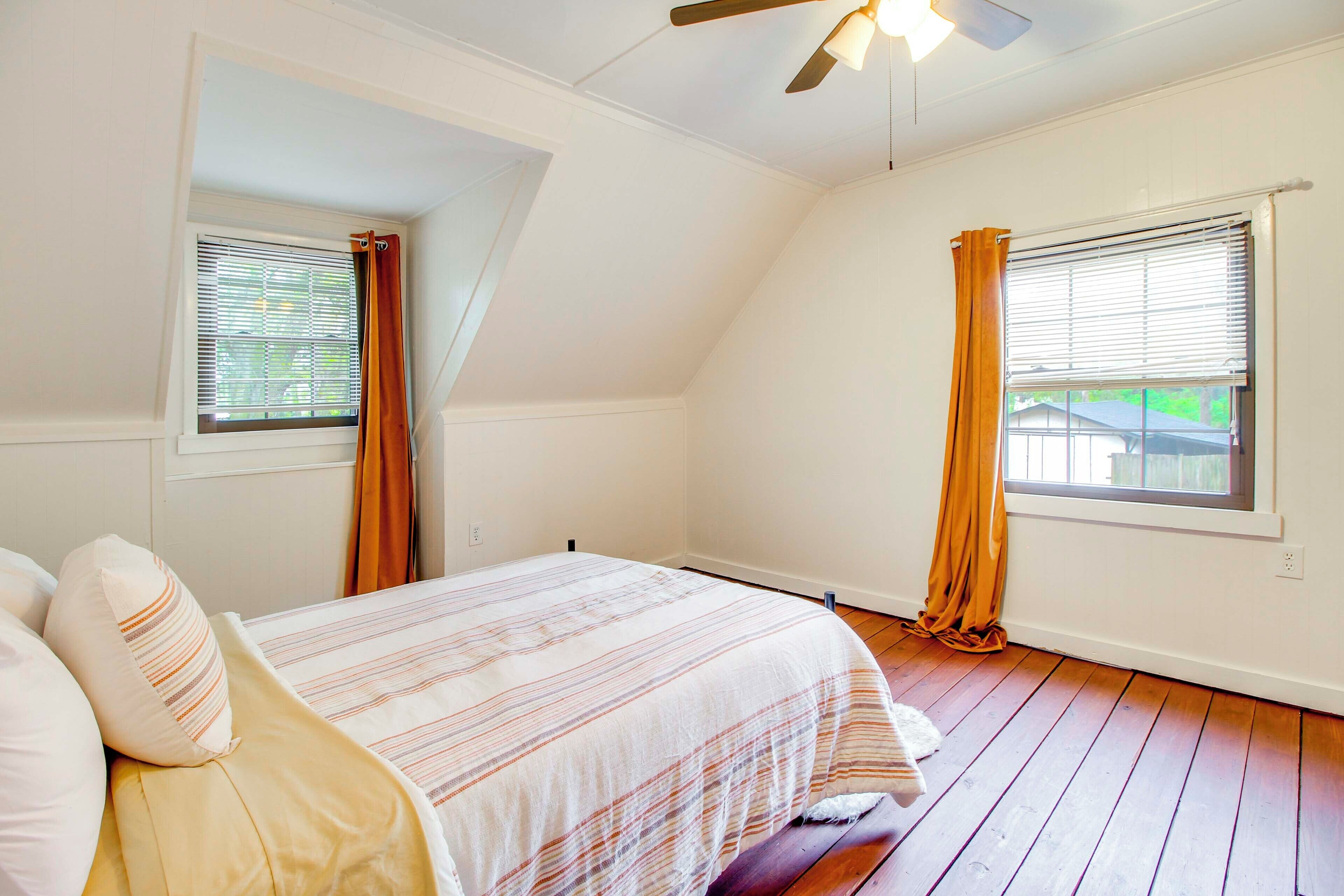 Un dormitorio con cama, dos ventanas con cortinas, un ventilador de techo y techo inclinado.