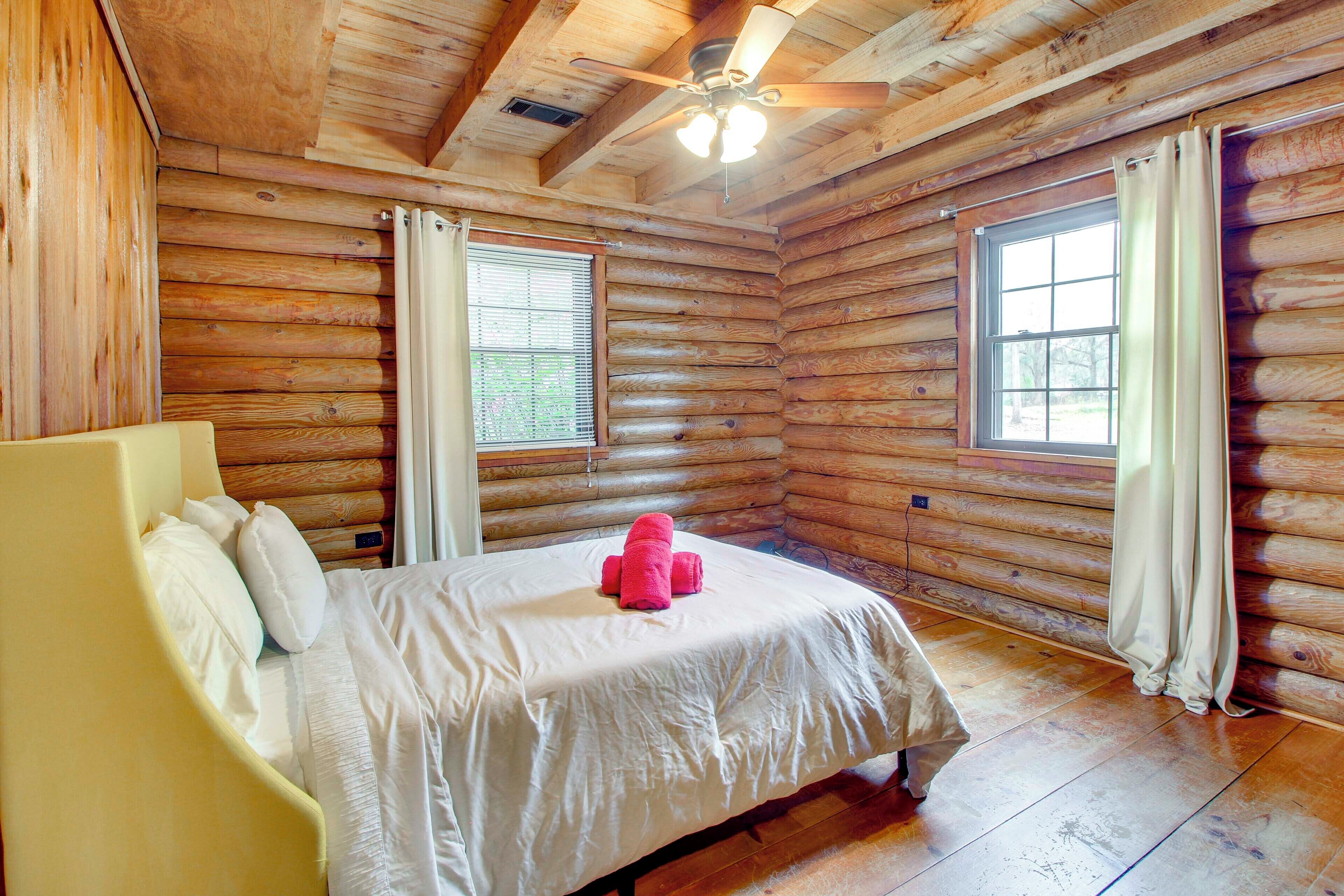 Ein Schlafzimmer in einer Blockhütte mit einem Bett, einem Deckenventilator und zwei mit Vorhängen versehenen Fenstern.