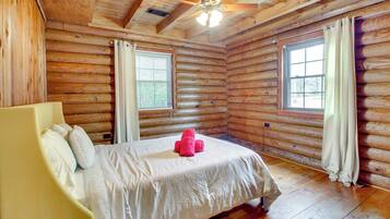 A log cabin bedroom with a bed, a ceiling fan, and two windows with curtains.