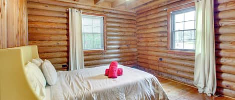 Ein Schlafzimmer in einer Blockhütte mit einem Bett, einem Deckenventilator und zwei mit Vorhängen versehenen Fenstern.