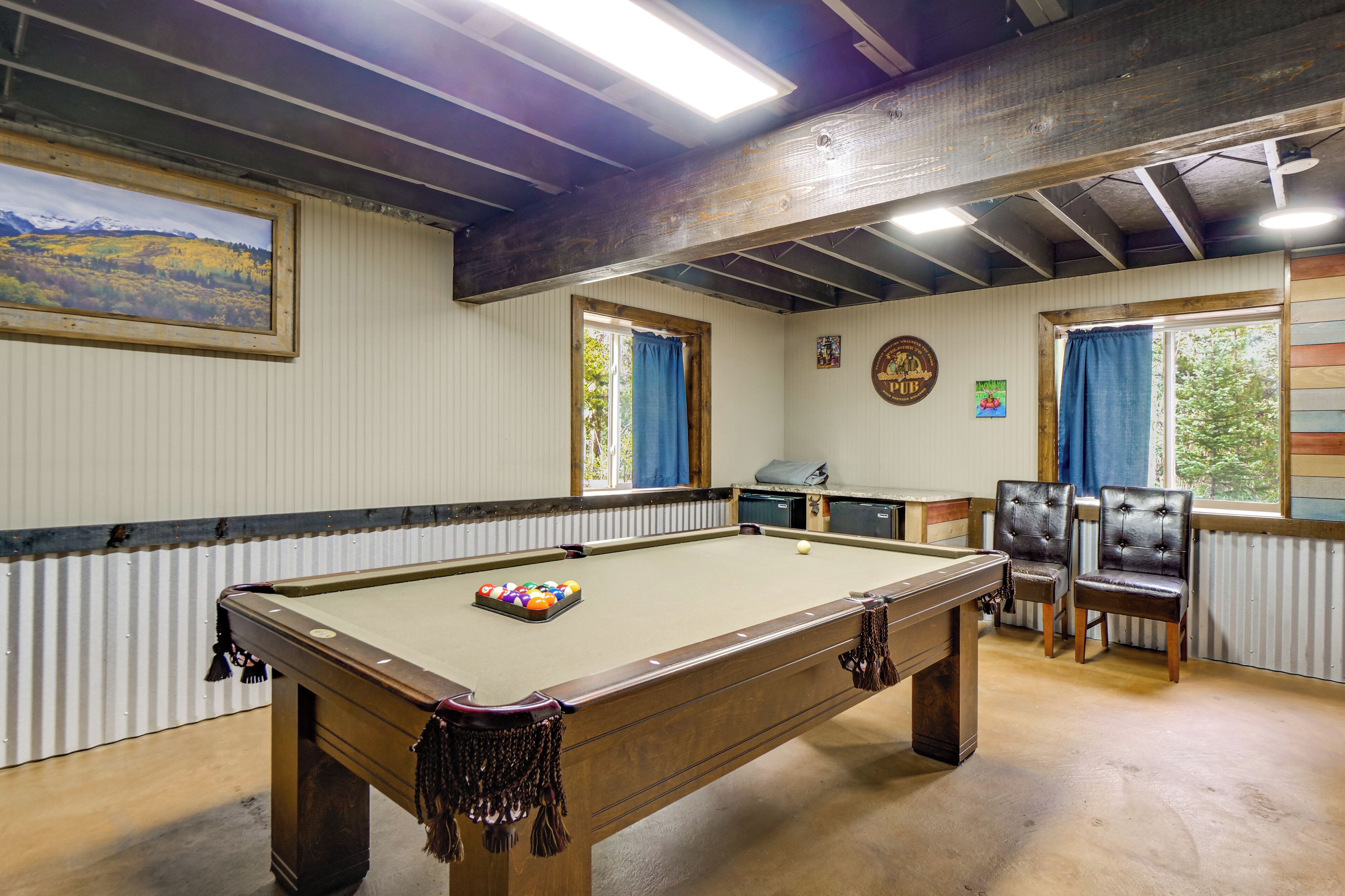 Een pooltafel met ballen, een poolkeu en een stoel. Er is een raam met gordijnen, een schilderij aan de muur en een klok.