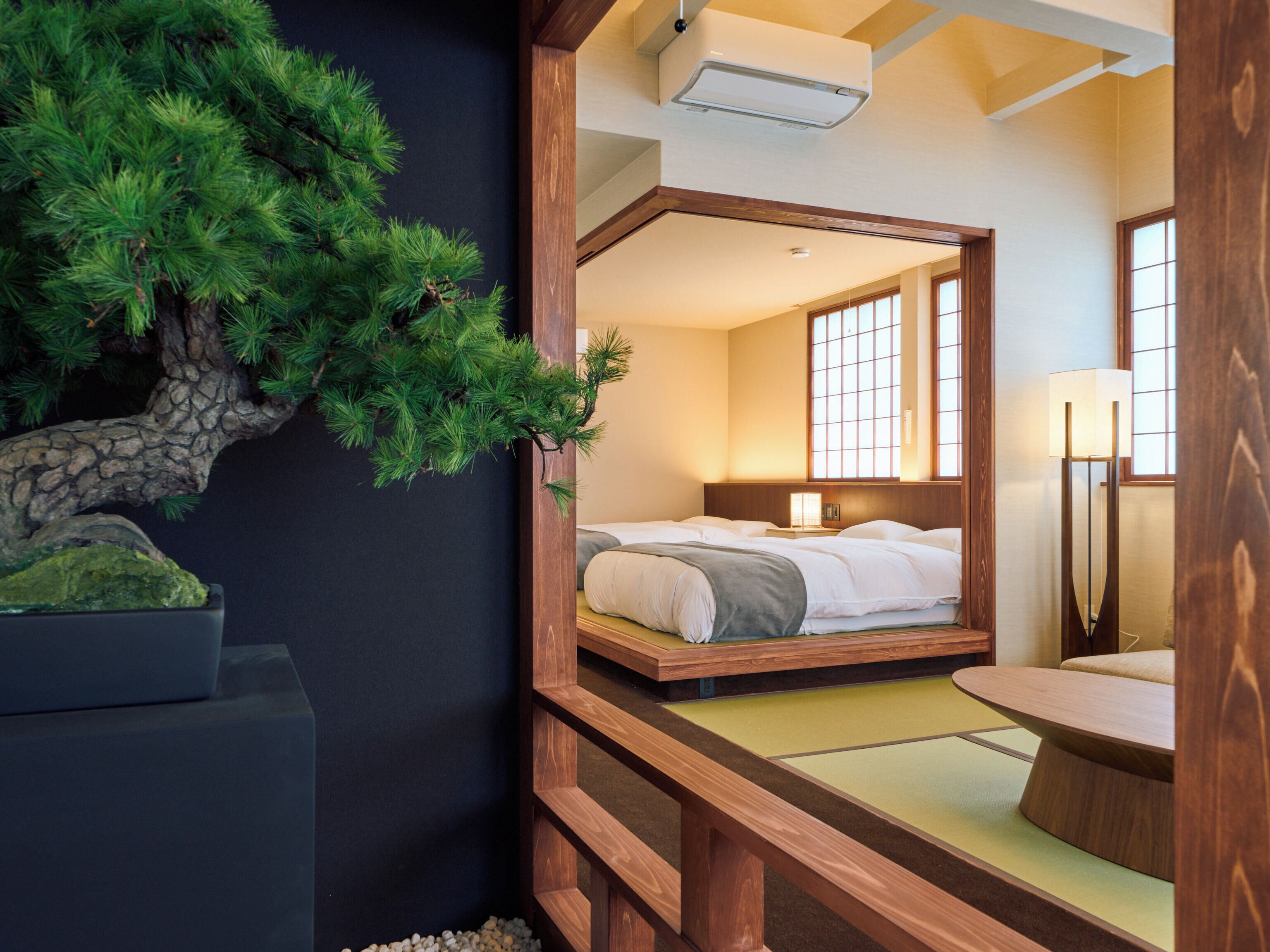A traditional Japanese-style room with tatami flooring, a low wooden bed, a small table, and a pine tree decoration.