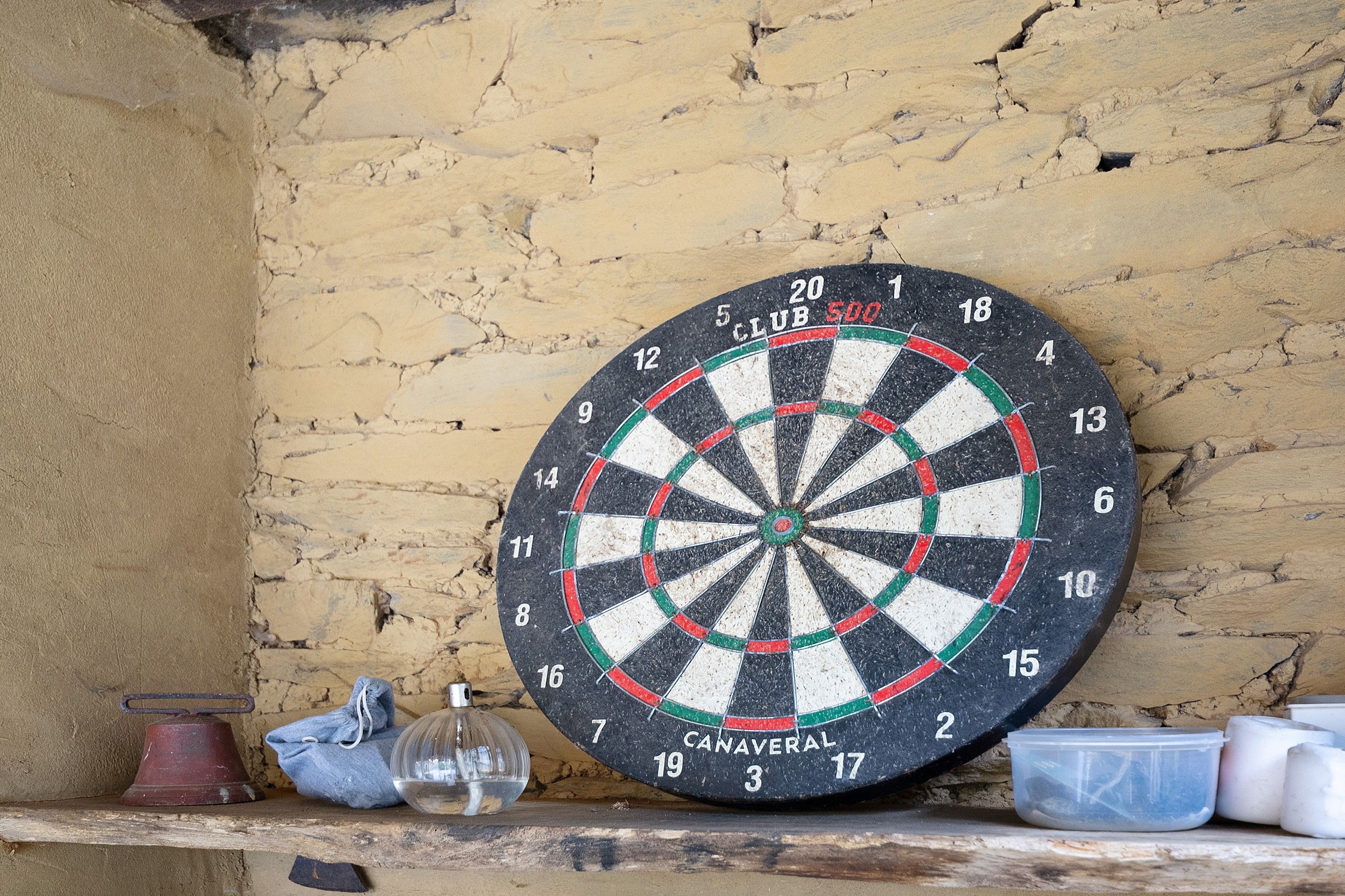 A dartboard mounted on a textured wall with a shelf below containing a bell, a glass, and plastic containers.