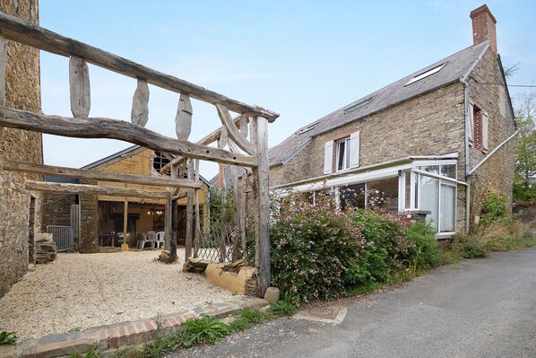 A rustic stone building with a wooden gate, a gravel driveway, and a small garden with pink flowers.
