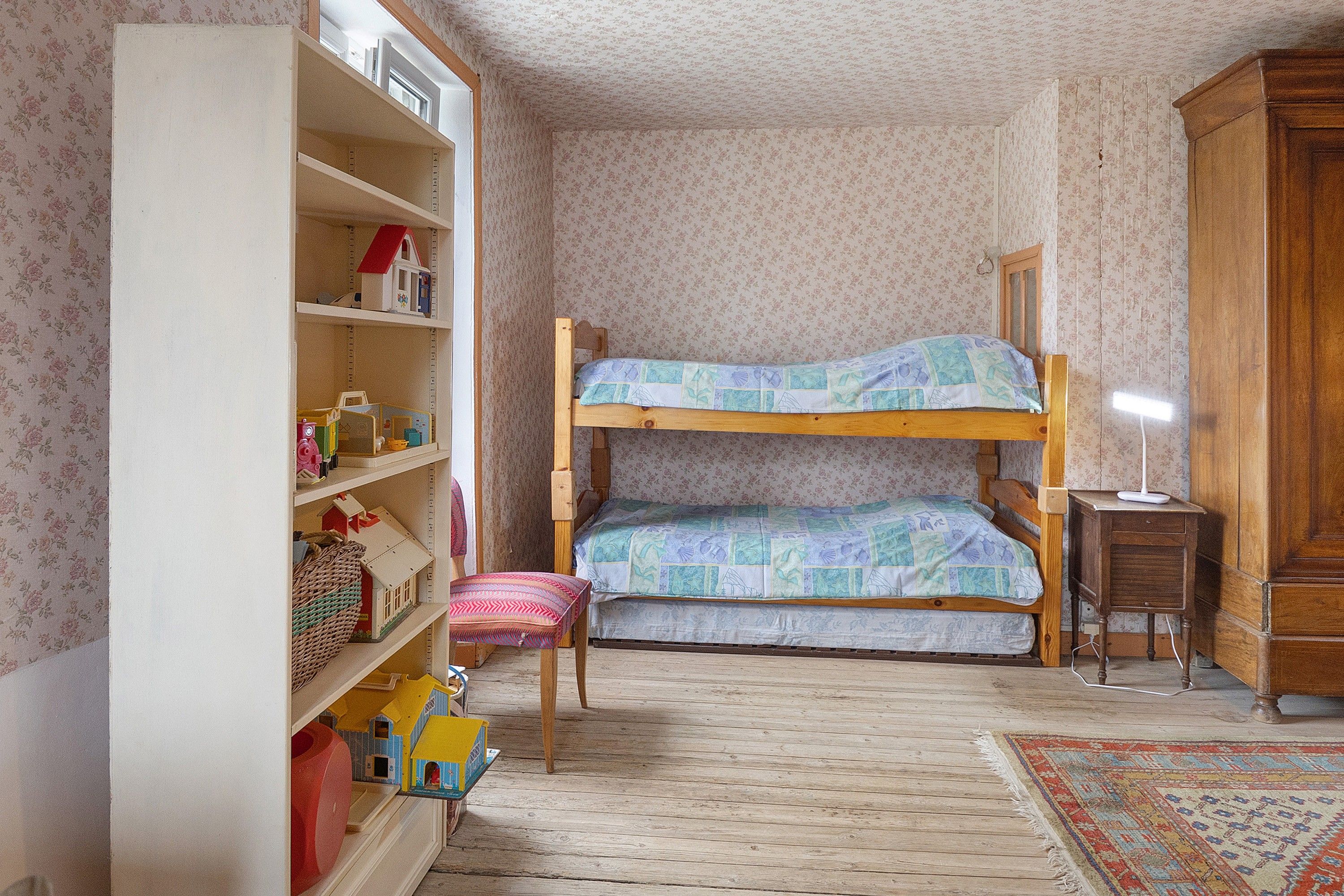 Une chambre d’enfant avec un lit superposé, une armoire en bois, un petit bureau et une bibliothèque remplie de jouets et de livres.