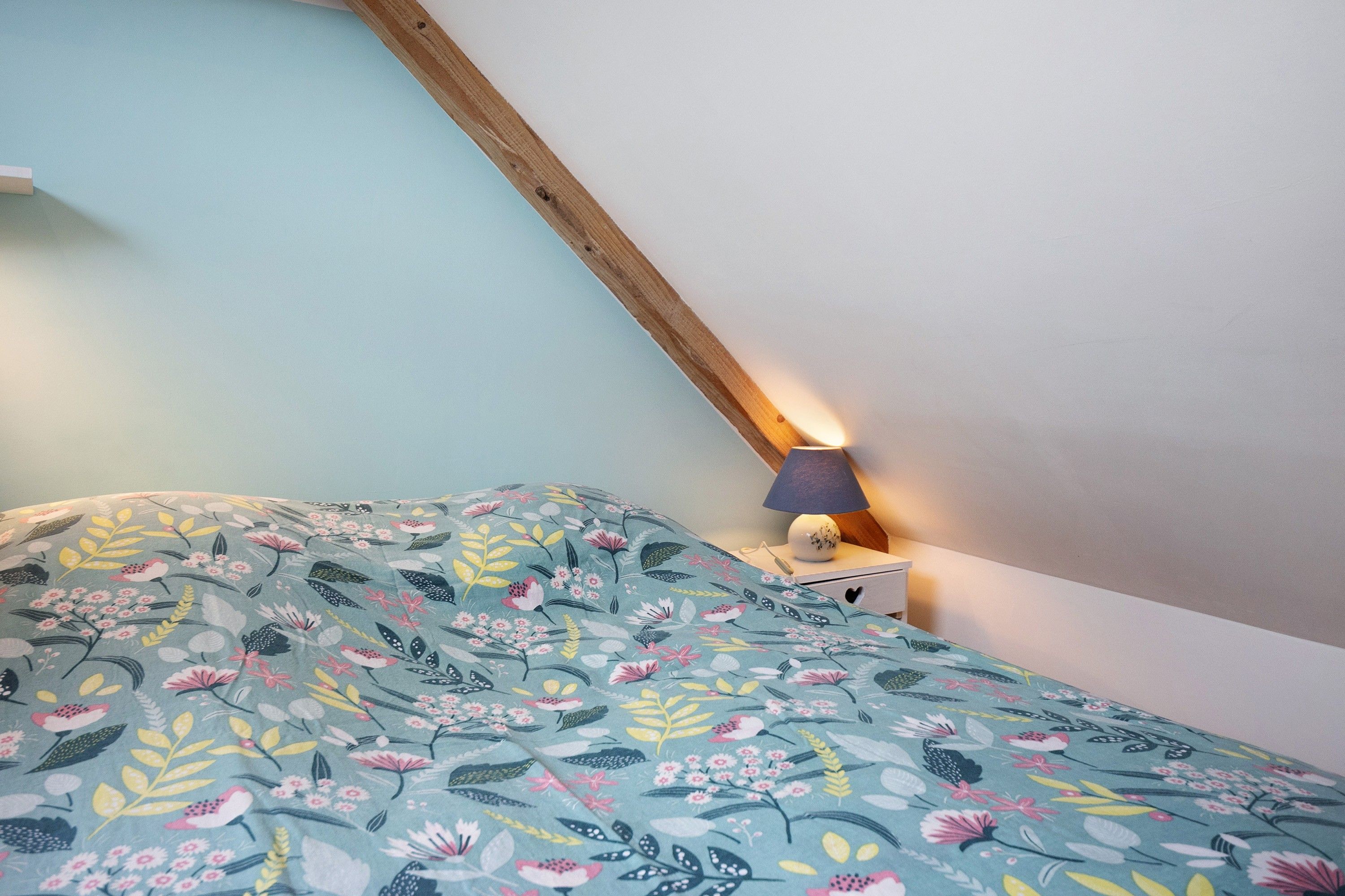 Une chambre avec un plafond en pente, un lit recouvert d’une couvre-lit à motifs floraux, une lampe de chevet et une table de chevet.