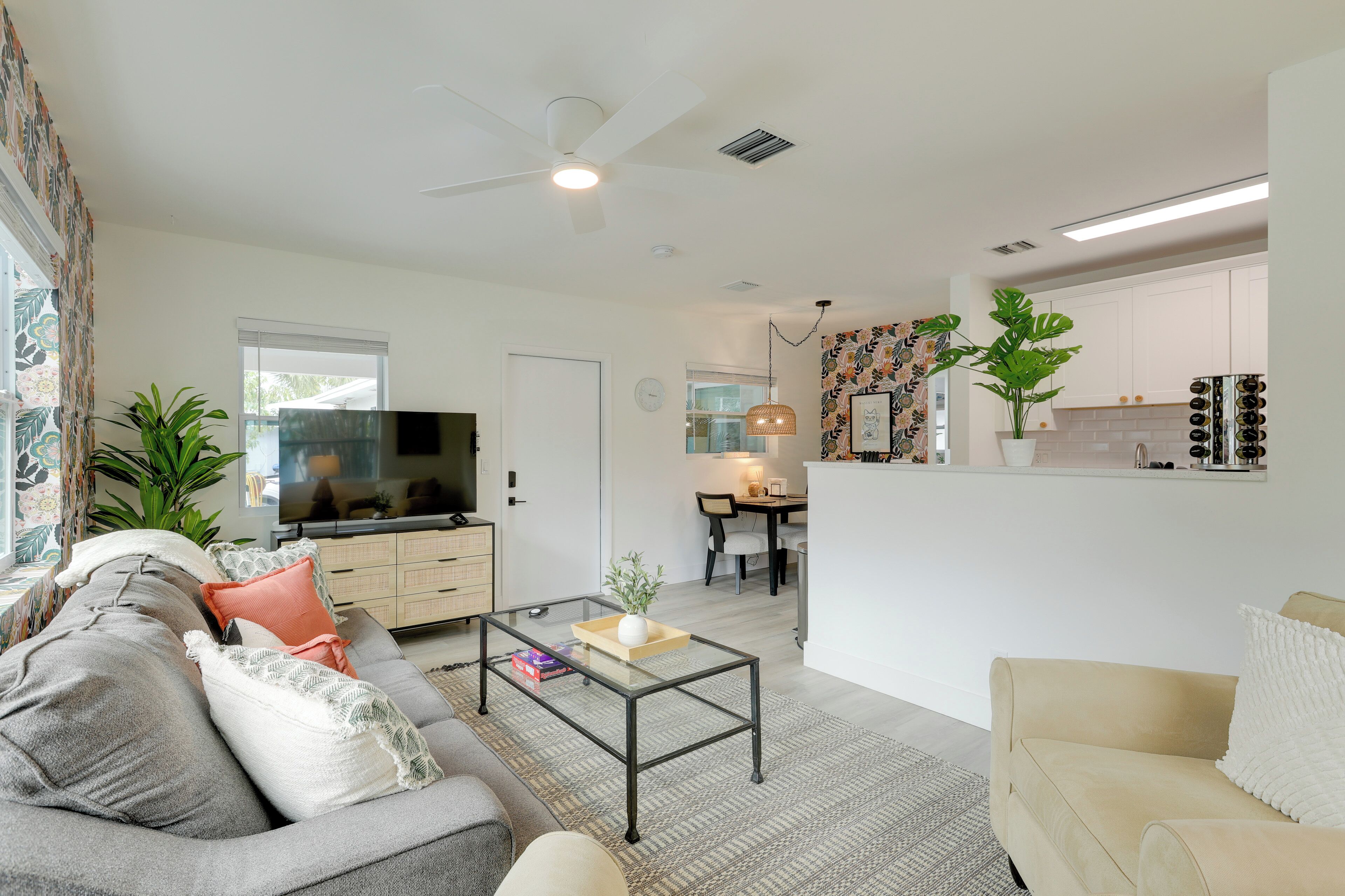A modern living room with a sofa, coffee table, and television. There is a kitchen area in the background with white cabinets and a dining table.