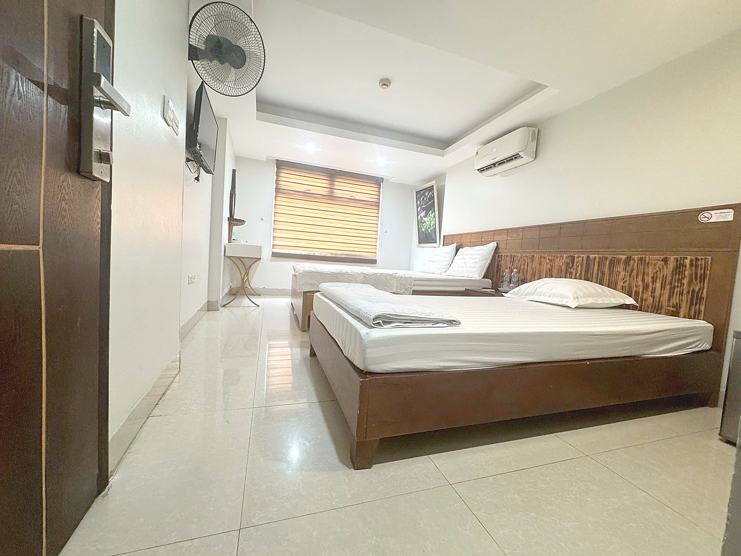 Une chambre d’hôtel avec un lit, une table de chevet, un ventilateur et une fenêtre avec des stores.