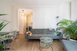 A modern living room with a grey sofa, a glass coffee table, and a dining area with a metal chair and table. - Charming and Bright 1BDR Near the Marais (Paris)