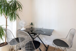Ein Eckzimmer mit Schreibtisch, zwei Drahtstuhle, einem Standventilator, einem kleinen Tisch und einer Topfpflanze.