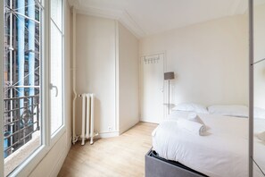 A hotel room with a bed, a radiator, a window with metal bars, and a door. - Charming and Bright 1BDR Near the Marais (Paris)