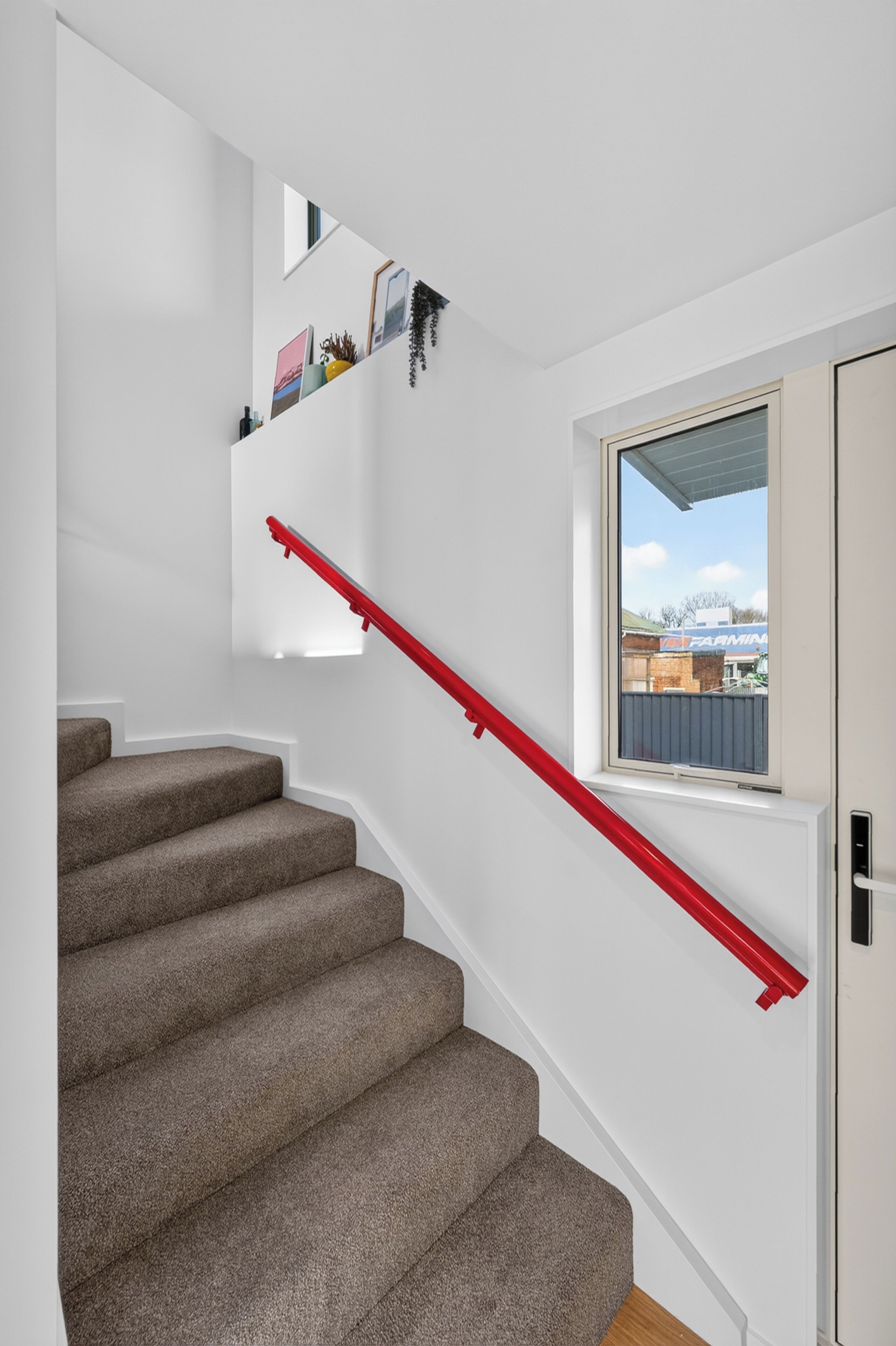 A staircase with carpeted steps and a red handrail, leading to a landing with a window overlooking a building.